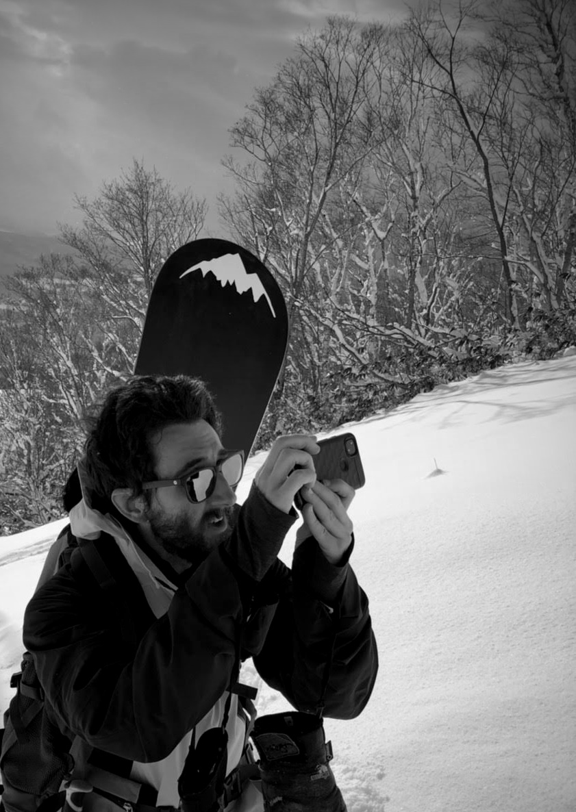 black and white photo of Samuel taking a photo while hiking with a snowboard.